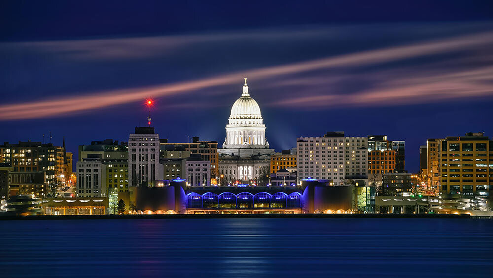 Nighttime view of downtown Madison, Wisconsin, featuring the illuminated Wisconsin State Capitol building reflected over Lake Monona.