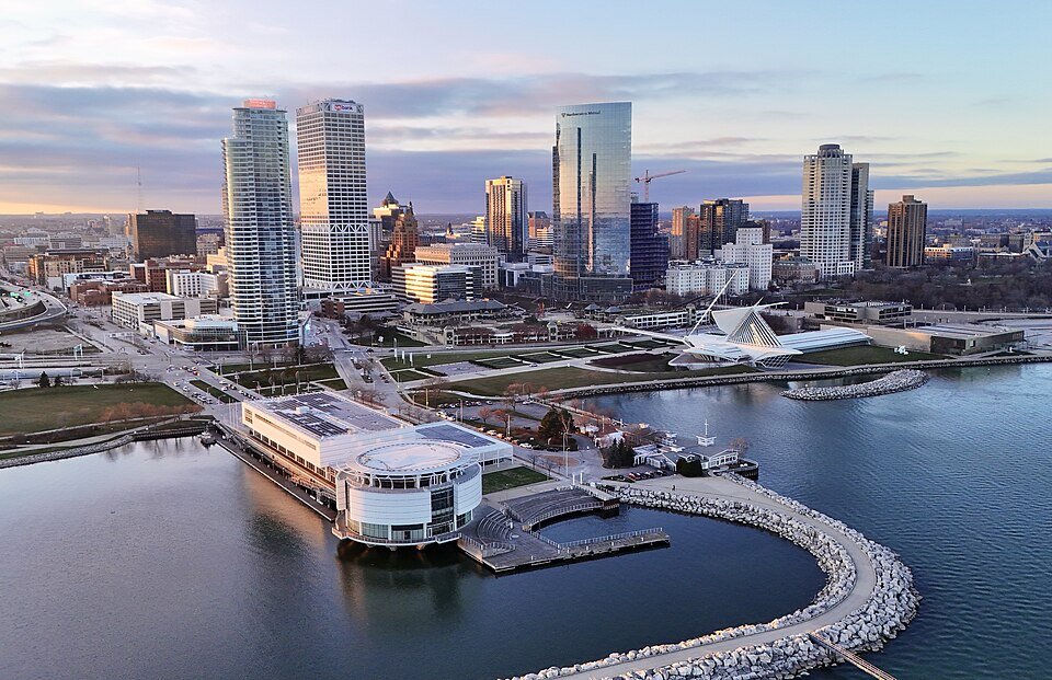 Aerial view of downtown Milwaukee skyline at sunset, featuring modern high-rise buildings, the Milwaukee Art Museum, and Lake Michigan shoreline.