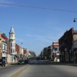 Historic downtown street view in Berlin, Wisconsin, featuring vintage brick buildings, a clock tower, American flags, and light traffic under a clear blue sky.