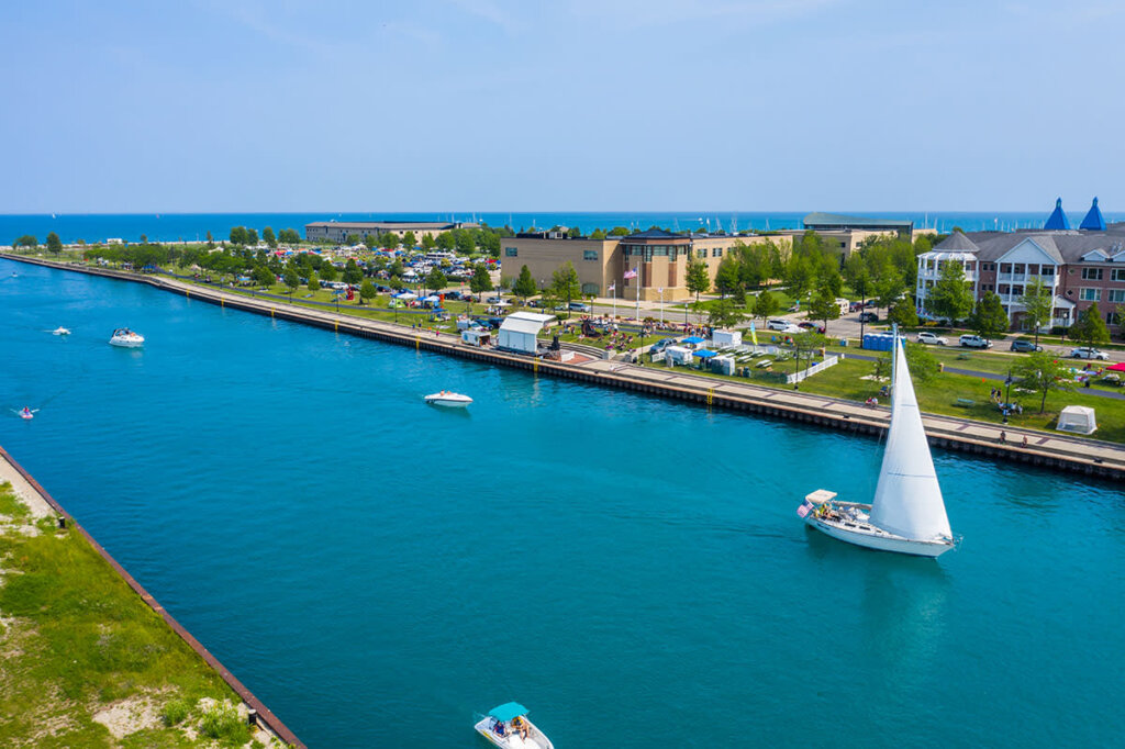 Boats and a sailboat glide along the bright blue water of Kenosha's scenic lakefront, with green parks, buildings, and Lake Michigan in the background.