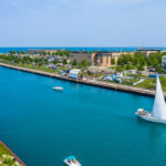 Boats and a sailboat glide along the bright blue water of Kenosha's scenic lakefront, with green parks, buildings, and Lake Michigan in the background.