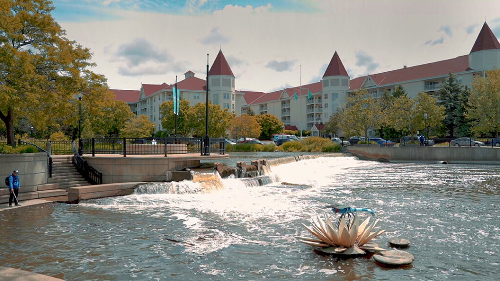 Scenic view of a charming senior living community in Waukesha, featuring a small waterfall, walking paths, and castle-like buildings with red roofs in the background.