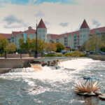Scenic view of a charming senior living community in Waukesha, featuring a small waterfall, walking paths, and castle-like buildings with red roofs in the background.