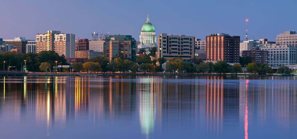 Evening skyline of downtown Madison, Wisconsin, with the State Capitol dome lit up and colorful city lights reflecting on the calm lake.