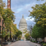 Scenic view of State Street in Madison, Wisconsin, lined with trees and historic buildings, leading to the Wisconsin State Capitol dome in the background.