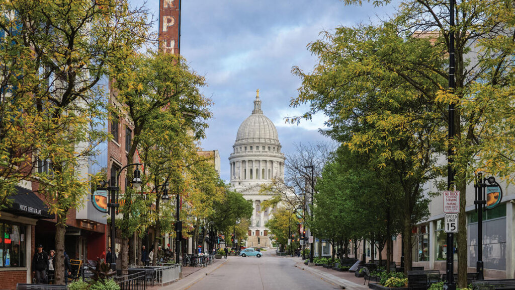 View of State Street in Madison, Wisconsin, lined with trees and shops, leading toward the Wisconsin State Capitol building under a cloudy sky.
