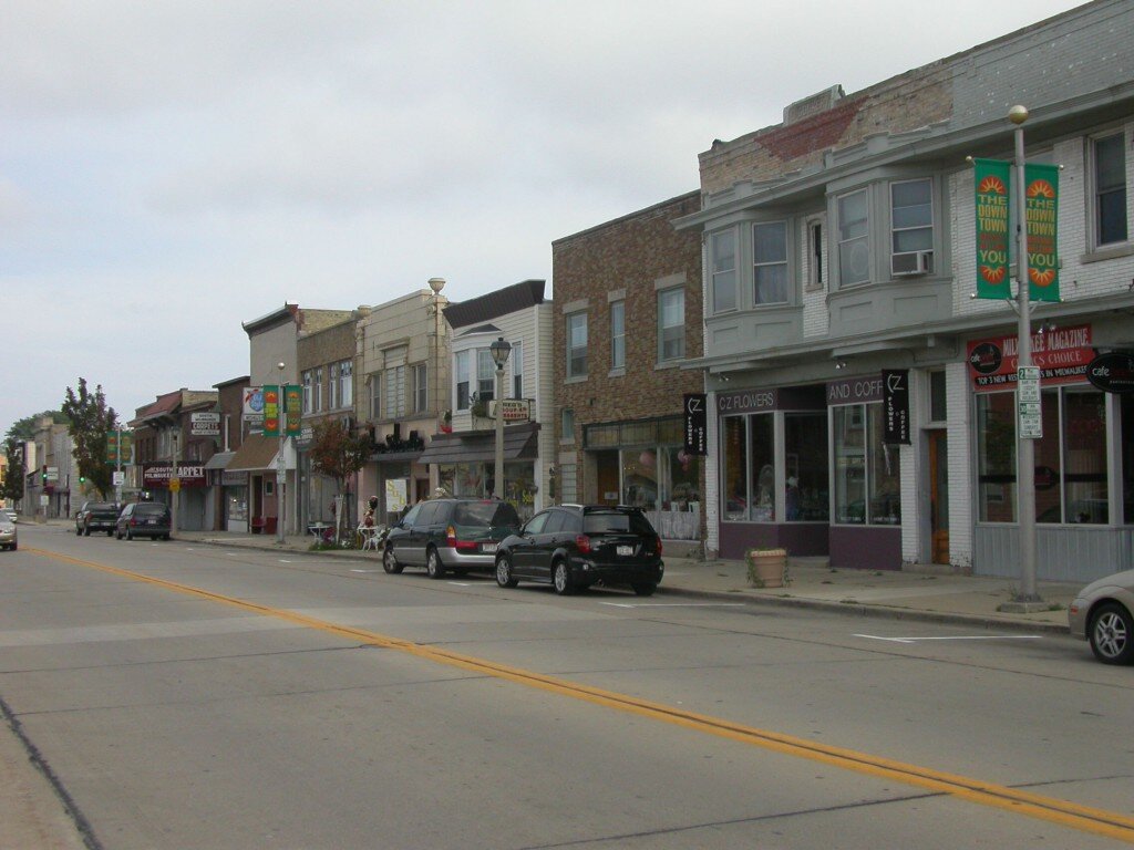 Downtown street view in West Allis, Wisconsin, featuring local shops, parked cars, and overcast skies.