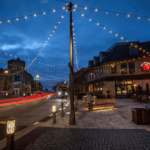 Charming evening street scene in Wauwatosa with string lights overhead, cozy outdoor seating, historic buildings, and light trails from passing cars.