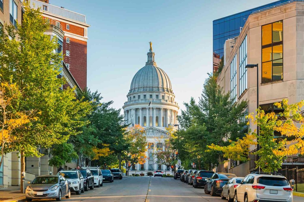 View of the Wisconsin State Capitol building in Madison, framed by tree-lined streets and parked cars on a sunny day.
