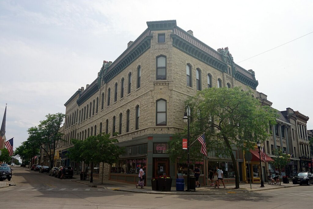 Historic stone building on a street corner in downtown Waukesha, Wisconsin, with American flags, storefronts, and pedestrians walking along the sidewalk.