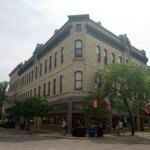 Historic stone building on a street corner in downtown Waukesha, Wisconsin, with American flags, storefronts, and pedestrians walking along the sidewalk.