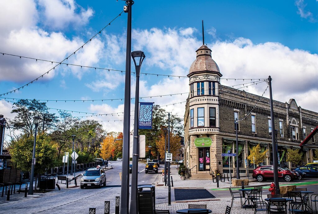 Historic building with turret in Wauwatosa Village, lined with shops, string lights, and autumn trees under a bright blue sky.