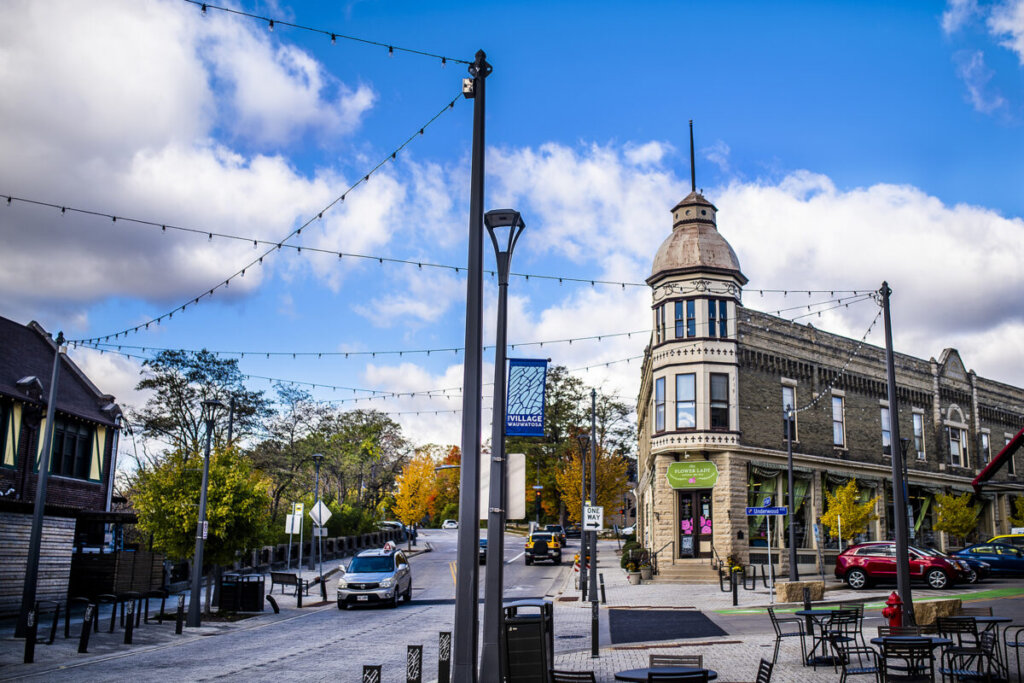 Charming street view of downtown Wauwatosa, featuring historic architecture, string lights, colorful trees, and local shops under a bright blue sky.