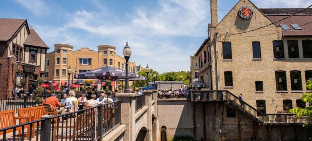 People dining outdoors on a sunny day in downtown Wauwatosa, surrounded by historic brick buildings and a small pedestrian bridge.