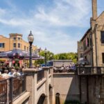 People dining outdoors on a sunny day in downtown Wauwatosa, surrounded by historic brick buildings and a small pedestrian bridge.
