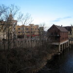Downtown Wauwatosa buildings along the Menomonee River on a clear day, with leafless trees in the foreground.
