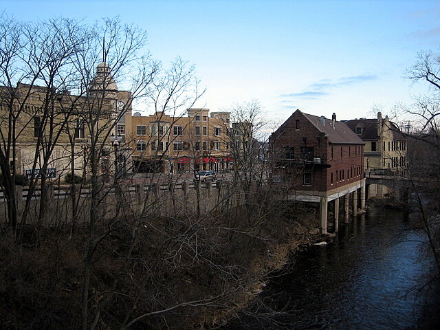 Downtown Wauwatosa buildings along the Menomonee River on a clear day, with leafless trees in the foreground.
