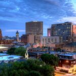 Downtown West Allis skyline at dusk with illuminated buildings and a colorful evening sky.