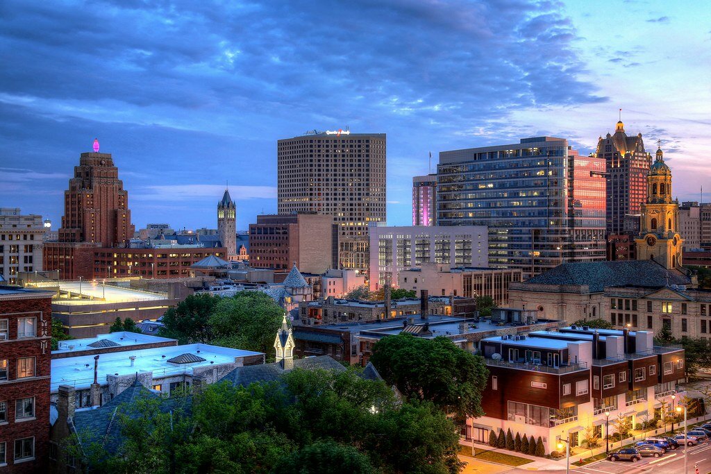 Downtown West Allis skyline at dusk with illuminated buildings and a colorful evening sky.