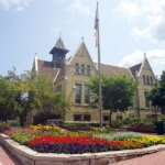 Historic brick building in West Allis surrounded by colorful flower beds, trees, and a flagpole under a partly cloudy sky.