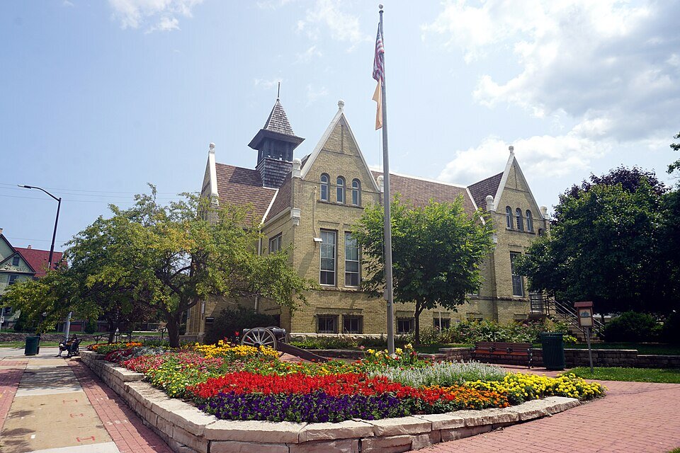 Historic brick building in West Allis surrounded by colorful flower beds, trees, and a flagpole under a partly cloudy sky.