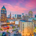 Downtown Milwaukee skyline at sunset with historic and modern buildings lit up under a vibrant pink and purple sky.
