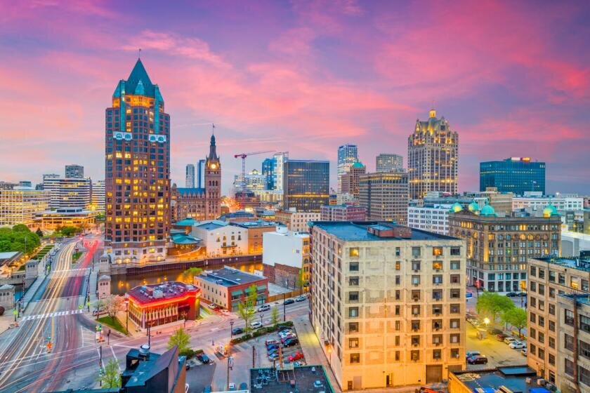 Downtown Milwaukee skyline at sunset with historic and modern buildings lit up under a vibrant pink and purple sky.