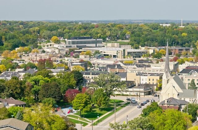Aerial view of downtown Waukesha, Wisconsin, showcasing residential neighborhoods, historic buildings, and a prominent church steeple surrounded by lush green trees.