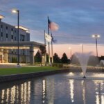 Exterior view of Hampton Inn & Suites Kenosha with a lit water fountain and flags at sunset.