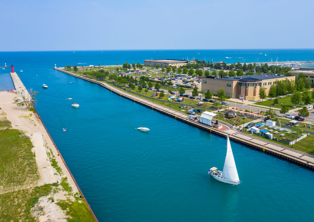 Aerial view of Kenosha Harbor with boats sailing through the bright blue water, surrounded by green parkland, modern buildings, and a bustling shoreline.