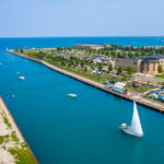 Aerial view of Kenosha Harbor with boats sailing through the bright blue water, surrounded by green parkland, modern buildings, and a bustling shoreline.