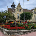 Historic brick building with pointed gables behind a vibrant garden of red, yellow, and pink flowers in West Allis, Wisconsin.
