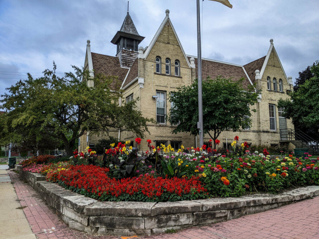 Historic brick building with pointed gables behind a vibrant garden of red, yellow, and pink flowers in West Allis, Wisconsin.