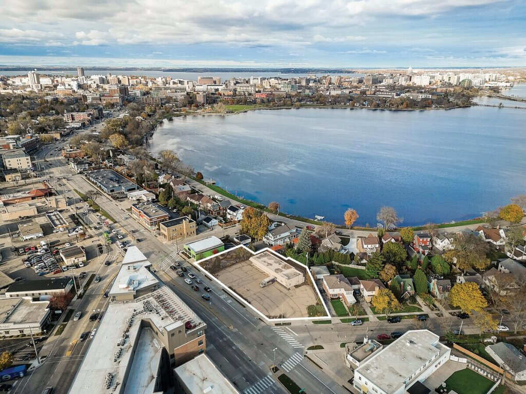 Aerial view of downtown Madison, Wisconsin, showing a mix of residential and commercial buildings near Lake Monona under a partly cloudy sky.