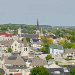 Aerial view of downtown Waukesha, Wisconsin, featuring historic churches, residential buildings, and lush greenery under a clear sky.