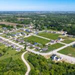 Aerial view of a residential neighborhood in Kenosha, Wisconsin, featuring new single-family homes, open green lots, and surrounding wooded areas under a clear blue sky.