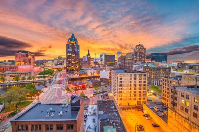 Downtown Milwaukee skyline at sunset with colorful clouds and lit buildings, showcasing the city's vibrant urban atmosphere.
