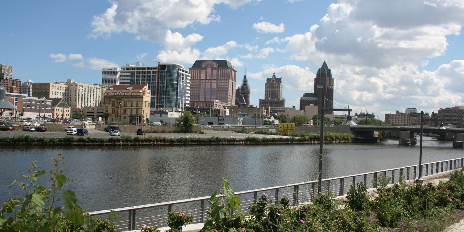 Downtown West Allis skyline with modern and historic buildings viewed across a calm river under a partly cloudy sky.