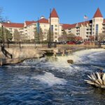 Riverwalk with small waterfall in downtown Waukesha, Wisconsin, featuring a decorative sculpture and large red-roofed buildings in the background.