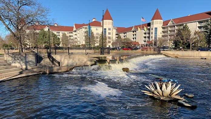 Riverwalk with small waterfall in downtown Waukesha, Wisconsin, featuring a decorative sculpture and large red-roofed buildings in the background.