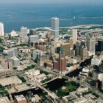Aerial view of downtown Milwaukee with tall buildings, highways, and Lake Michigan in the background.