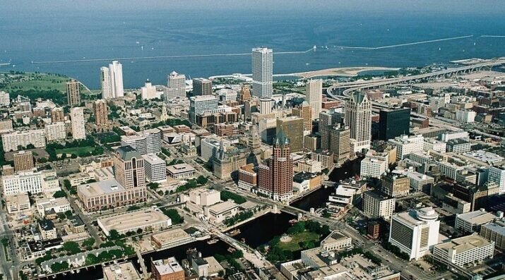 Aerial view of downtown Milwaukee with tall buildings, highways, and Lake Michigan in the background.