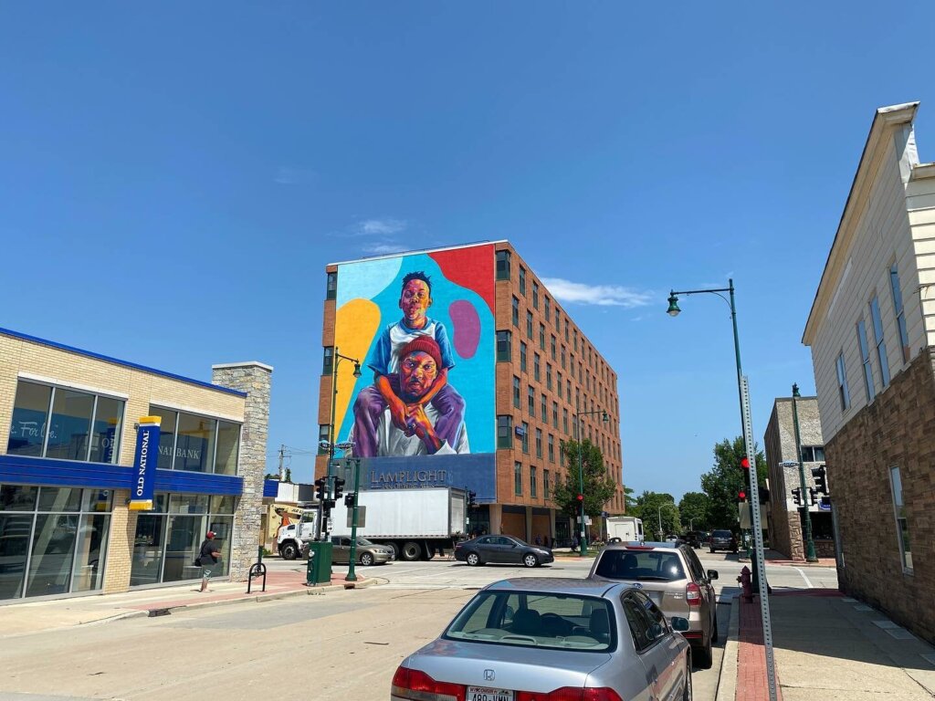 Colorful mural of two people on the side of a brick building in downtown West Allis, with cars parked along the street and clear blue skies above.
