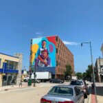 Colorful mural of two people on the side of a brick building in downtown West Allis, with cars parked along the street and clear blue skies above.
