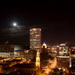 Milwaukee city skyline at night with illuminated buildings and a full moon over Lake Michigan.