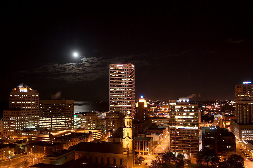 Milwaukee city skyline at night with illuminated buildings and a full moon over Lake Michigan.
