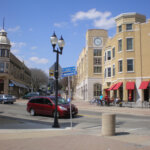 Downtown Wauwatosa intersection featuring historic and modern buildings, a red minivan, pedestrians, and clear street signs under a blue sky.