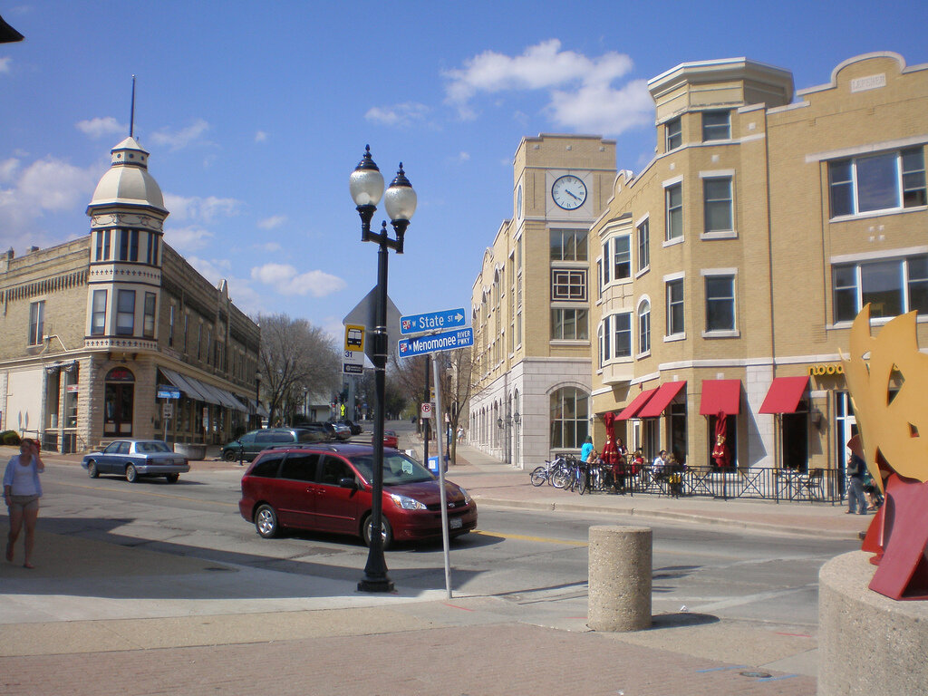 Downtown Wauwatosa intersection featuring historic and modern buildings, a red minivan, pedestrians, and clear street signs under a blue sky.