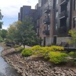Modern brick apartment buildings alongside a rocky riverbank with trees and shrubs in Waukesha.