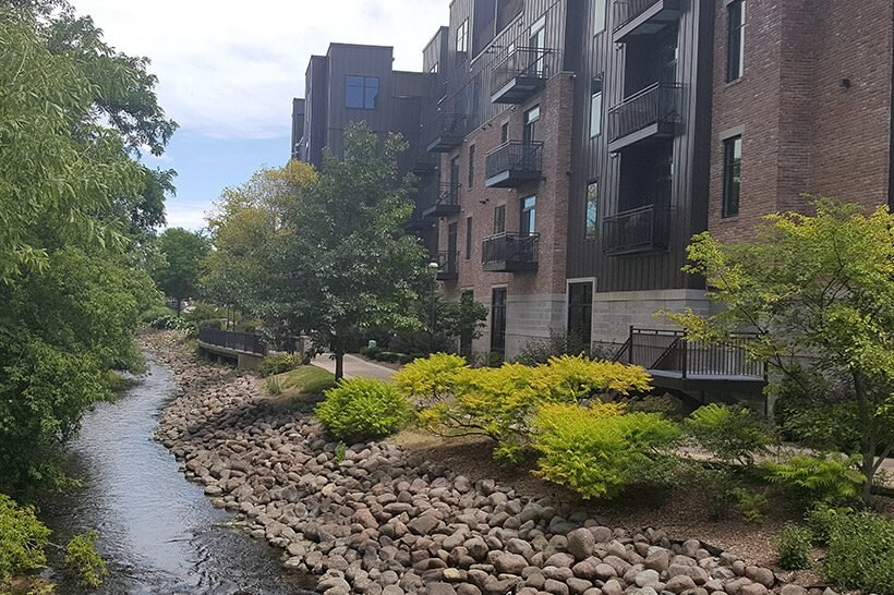 Modern brick apartment buildings alongside a rocky riverbank with trees and shrubs in Waukesha.